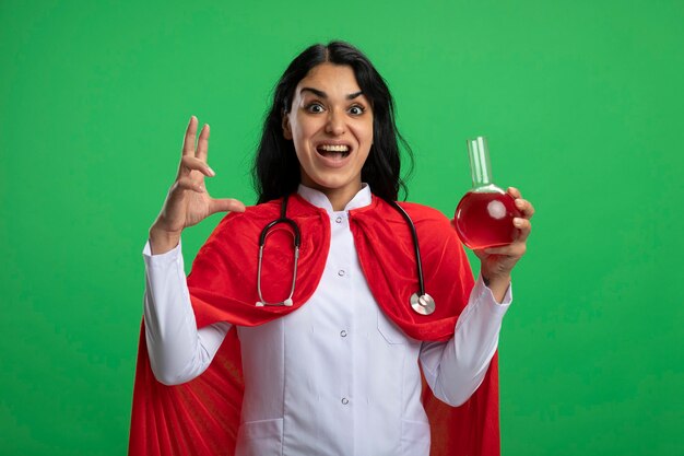 Emocionada joven superhéroe vestida con bata médica con estetoscopio sosteniendo una botella de vidrio de química llena de líquido rojo y mostrando el tamaño aislado en verde