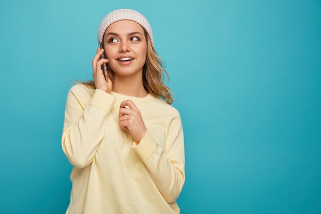 Emocionada joven vistiendo gorro de invierno hablando por teléfono mirando hacia arriba