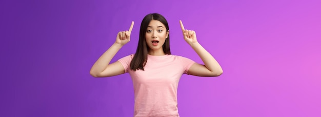 Emocionada linda chica asiática sorprendida en camiseta rosa contando noticias increíbles señalando con el dedo levantado