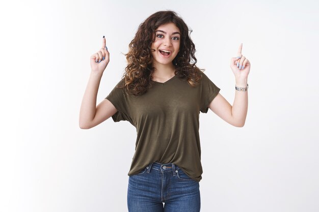 Emocionada, linda, energizada, joven y creativa georgiana con cabello rizado sonriendo le anima a probar el producto apuntando hacia arriba con los dedos índices hacia arriba promoviendo publicidad fresca, fondo blanco.