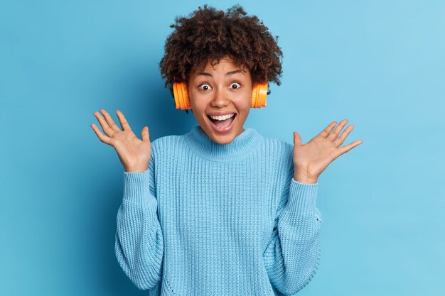 Emocionada mujer afroamericana se divierte en el interior levanta las palmas de las manos y exclama con alegría escucha su música favorita a través de auriculares estéreo y usa poses de suéter informal