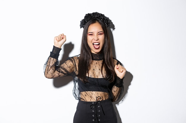 Emocionada mujer asiática en elegante vestido de encaje gótico y corona celebrando halloween, disfrutando de la fiesta, levantando la mano y gritando alegre, de pie sobre fondo blanco.