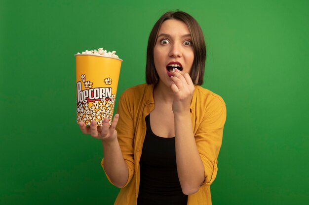 Emocionada mujer bonita caucásica come y sostiene un cubo de palomitas de maíz en verde