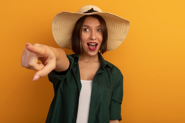 Emocionada mujer bonita con sombrero de playa se ve y apunta al lado aislado en la pared naranja