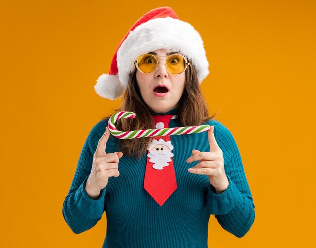 Emocionada mujer caucásica adulta en gafas de sol con gorro de Papá Noel y corbata de santa sosteniendo bastón de caramelo aislado en la pared naranja con espacio de copia