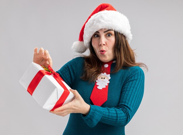 Emocionada mujer caucásica adulta con gorro de Papá Noel y corbata de santa con caja de regalo de navidad aislada en la pared blanca con espacio de copia