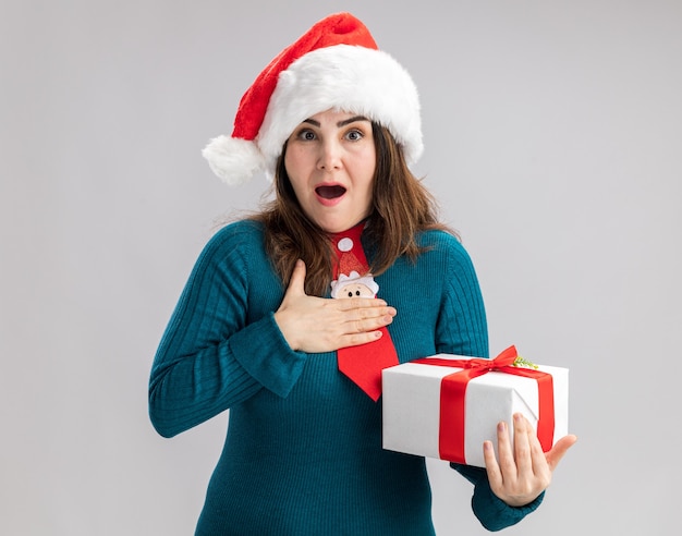 Emocionada mujer caucásica adulta con gorro de Papá Noel y corbata de santa pone la mano en el pecho y sostiene la caja de regalo de Navidad aislada sobre fondo blanco con espacio de copia