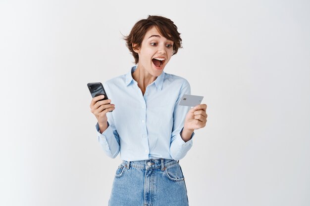 Emocionada mujer caucásica mirando tarjeta de crédito de plástico y sosteniendo el teléfono inteligente, de pie sobre la pared blanca