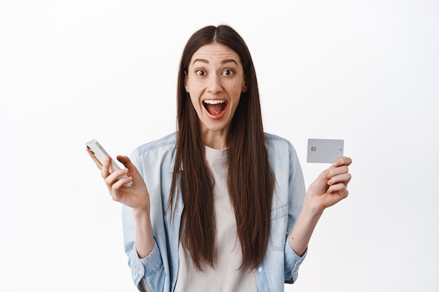 Emocionada mujer caucásica mostrando tarjeta de crédito y sosteniendo un teléfono inteligente, haciendo un pedido, esperando la entrega mientras compra en línea, de pie sobre una pared blanca