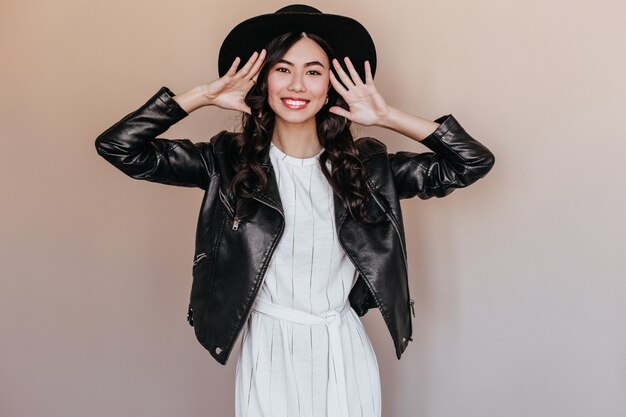 Emocionada mujer china con sombrero negro mirando a la cámara. Vista frontal del increíble modelo asiático en chaqueta de cuero posando con una sonrisa.