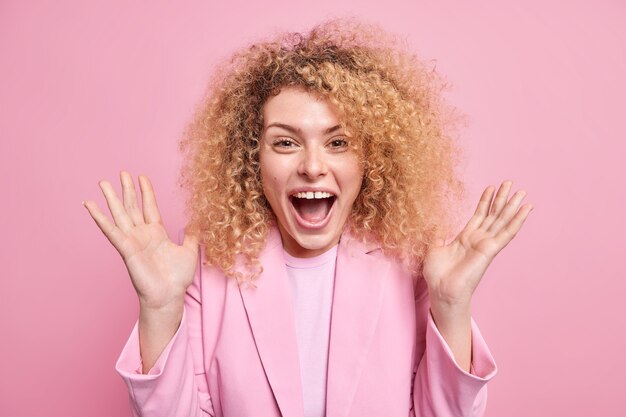 Emocionada mujer europea de pelo rizado levanta las palmas de las manos exclama en voz alta mantiene la boca abierta reacciona ante una noticia increíble