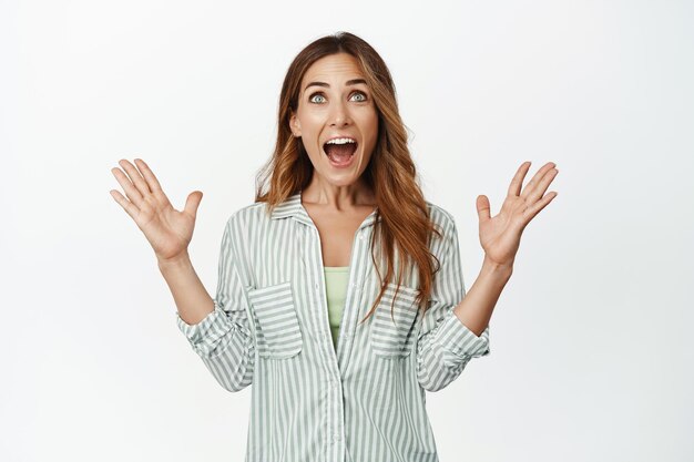 Emocionada mujer exitosa ganando, gritando de alegría y felicidad, dándose la mano con incredulidad, mirando hacia arriba en la clasificación del ganador, logrando la meta, celebrando, de pie contra el fondo blanco.