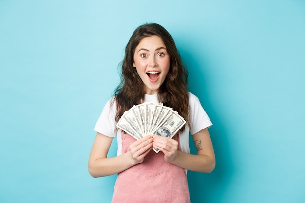 Emocionada mujer linda ganando dinero, sosteniendo billetes de un dólar y sonriendo asombrada, obtuvo crédito rápido, de pie sobre fondo azul.