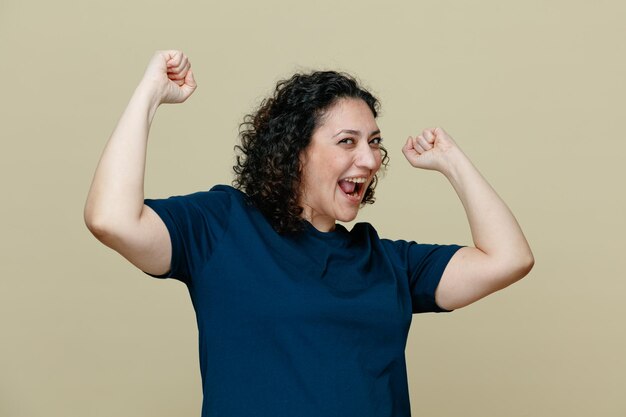 Emocionada mujer de mediana edad con camiseta mirando a la cámara levantando los puños mostrando un gesto de sí aislado en un fondo verde oliva