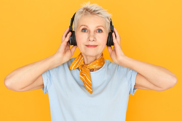 Emocionada mujer de mediana edad positiva con pelo corto teñido y ojos azules posando aislado en auriculares inalámbricos disfrutando de buena música