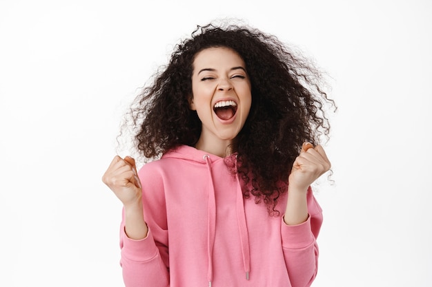 Emocionada mujer morena ganadora, lograr la meta o el éxito, gritando de alegría y placer, ganando un premio, celebrando la victoria con una expresión de alivio en blanco