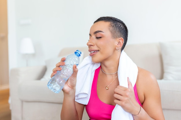 Emocionada mujer negra atlética en ropa deportiva riéndose sentada en una alfombra de yoga sosteniendo una botella de agua con una toalla en los hombros