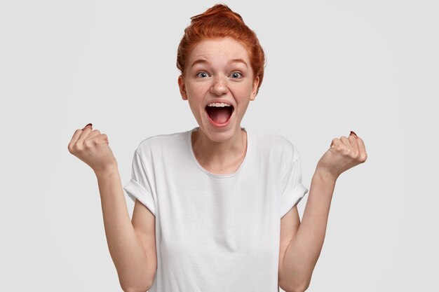 Emocionada mujer pecosa llena de alegría con cabello pelirrojo, levanta los puños y exclama en voz alta, anima a un amigo, grita palabras de apoyo, usa una camiseta blanca de maqueta informal, modelos y gestos