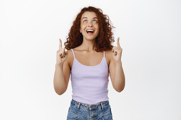 Emocionada mujer pelirroja sonriente, riendo y luciendo feliz, señalando con el dedo hacia arriba, mostrando publicidad promocional, de pie contra el fondo blanco