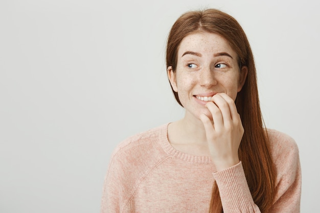 Emocionada mujer pelirroja sonriente toca el labio y mira a la izquierda con tentación