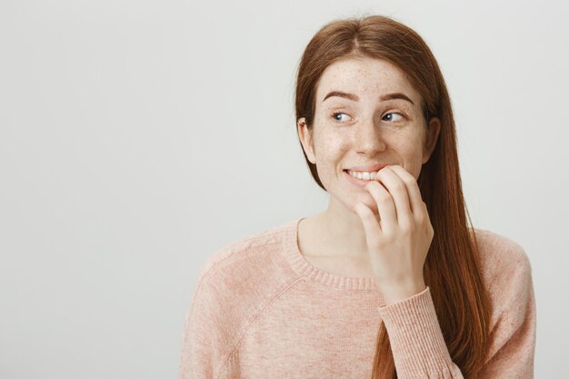 Emocionada mujer pelirroja sonriente toca el labio y mira a la izquierda con tentación