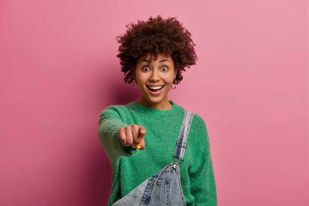 Emocionada mujer positiva tiene peinado afro, señala con el dedo índice directamente a usted, ve algo muy divertido, usa un suéter verde y un mono, aislado sobre una pared rosa pastel. Increible