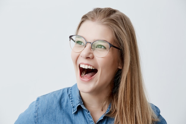 Emocionada mujer rubia con gafas, camisa de mezclilla, exclama alegremente, feliz de saber acerca de la inscripción a la universidad, sueña con ser estudiante por mucho tiempo, no puede creer en el éxito.