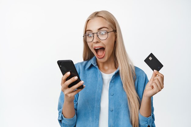 Emocionada mujer rubia con gafas mirando la pantalla del teléfono inteligente y gritando, mirando sorprendido y asombrado en blanco