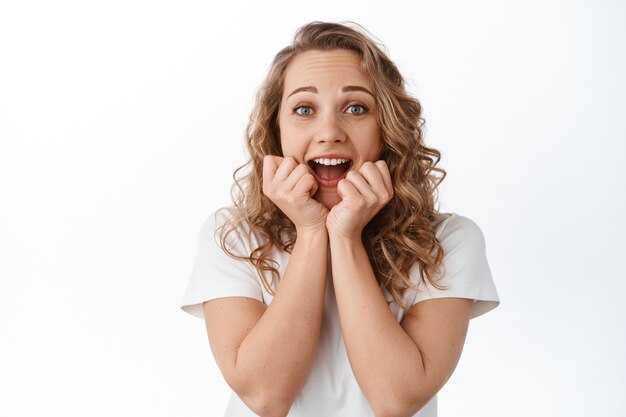 Emocionada mujer rubia grita de alegría y sorprendida, ganando y celebrando, gritando de felicidad y asombro, de pie con una camiseta sobre la pared blanca