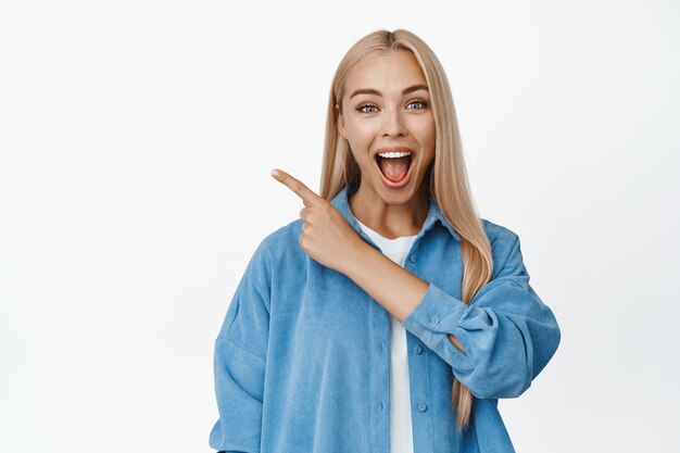 Emocionada mujer rubia sonriendo, señalando con el dedo en la esquina superior izquierda con cara de asombro, mostrando noticias increíbles, pancarta de gran venta en blanco