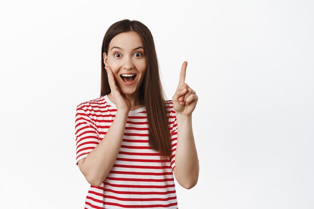 Emocionada mujer sonriente apuntando con el dedo hacia arriba, gesto de eureka, sugiriendo algo, tiene una idea, mostrando pancarta de venta, de pie contra la pared blanca