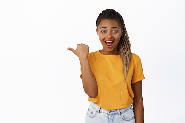 Emocionada niña africana sonriente apuntando a la izquierda y mirando asombrada al ver la promoción que demuestra la pancarta a un lado en el fondo blanco del espacio de copia