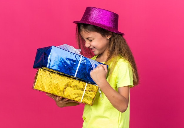 Foto gratuita emocionada niña caucásica con gorro de fiesta púrpura sosteniendo cajas de regalo y manteniendo el puño aislado en la pared rosa con espacio de copia