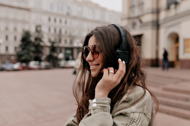 Emocionada niña feliz con cabello oscuro con chaqueta caqui y elegantes gafas escuchando música con auriculares y disfrutando de caminar por la calle en la ciudad vieja