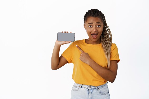 Emocionada niña negra sonriente señalando con el dedo a la pantalla del teléfono inteligente que muestra la interfaz de la aplicación del teléfono móvil recomendando el fondo blanco de la aplicación