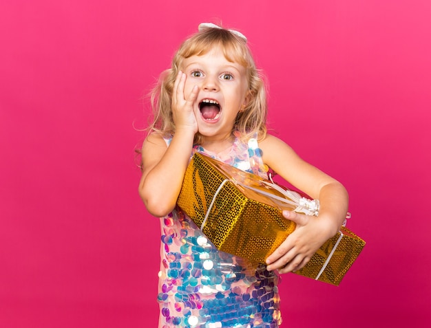Emocionada niña rubia poniendo la mano en la cara y sosteniendo la caja de regalo aislada en la pared rosa con espacio de copia
