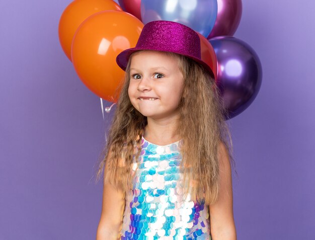 Emocionada niña rubia con sombrero de fiesta violeta de pie con globos de helio aislado en la pared púrpura con espacio de copia