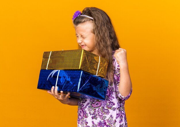 Emocionada niña rubia sosteniendo cajas de regalo y manteniendo el puño hacia arriba aislado en la pared naranja con espacio de copia