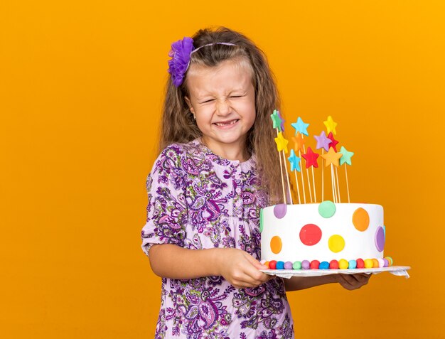 Emocionada niña rubia sosteniendo pastel de cumpleaños aislado en la pared naranja con espacio de copia