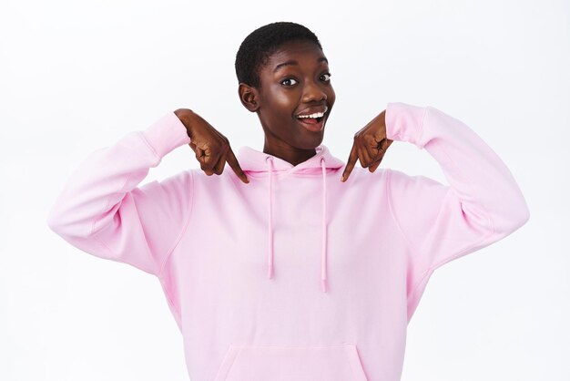 Emocionada y optimista mujer afroamericana amigable con el pelo corto en una sudadera con capucha rosa, apuntando con el dedo hacia abajo