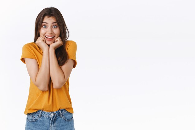 Emocionada, tonta y fascinada mujer joven adora ver un tráiler increíble con sus actores favoritos, tomados de la mano en la línea de la mandíbula y apretar las mejillas emocionada, sonriendo asombrada, de pie asombrado fondo blanco