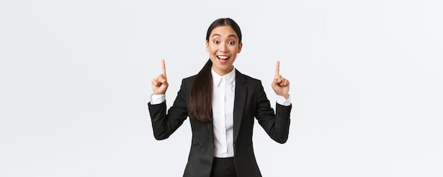 Emocionada vendedora asiática sonriente en traje sugiere mucho señalar con el dedo hacia arriba como detalles reveladores Empresaria haciendo un anuncio y mostrando el fondo blanco de la pancarta superior