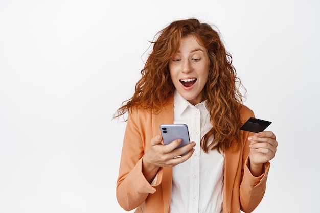 Emocionada vendedora pelirroja mirando la pantalla del teléfono móvil con la cara sorprendida ordenando compras en línea con teléfono inteligente y tarjeta de crédito de pie sobre fondo blanco
