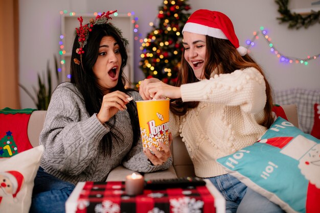 Emocionadas chicas jóvenes con gorro de Papá Noel y corona de acebo, sostén y miran el cubo de palomitas de maíz sentados en sillones y disfrutando de la Navidad en casa
