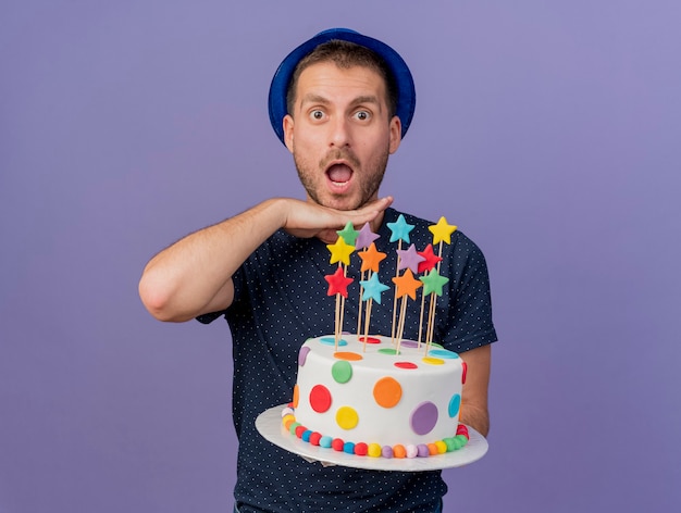 Emocionado apuesto hombre caucásico con sombrero azul mantiene la mano debajo de la barbilla y sostiene el pastel de cumpleaños aislado sobre fondo púrpura con espacio de copia