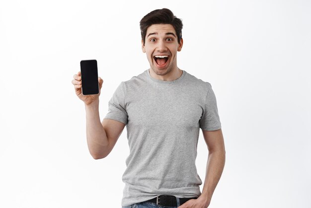 Emocionado chico feliz muestra la pantalla vacía del teléfono móvil haciendo publicidad en línea teléfono inteligente de pie contra el fondo blanco