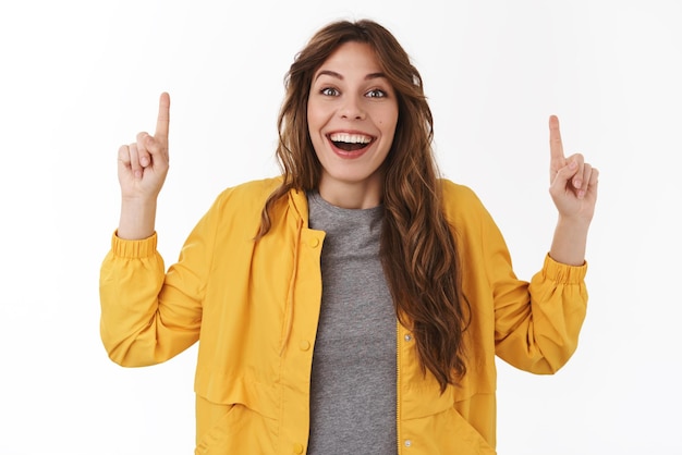 Emocionado emotivo feliz niña caucásica de 25 años mirada fascinada sonrisa entusiasta mirando a la cámara emocionado señalando hacia arriba diciendo oferta increíble promoción oportunidad de suerte de pie fondo blanco