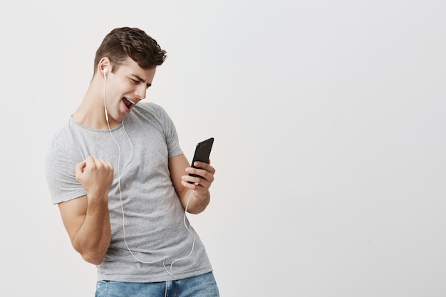 Emocionado ganador masculino levantando el puño con gran felicidad después de recibir un mensaje de su novia, mirando la pantalla de su teléfono inteligente. Joven campeón teniendo éxito. Concepto de victoria y triunfo