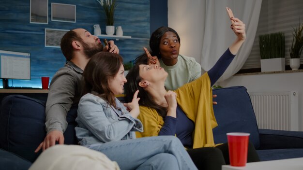 Emocionado grupo de amigos multirraciales sentados en el sofá durante la fiesta alegre tomando selfie