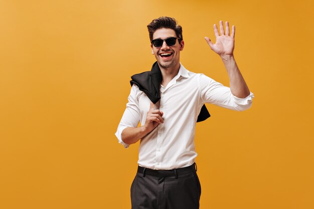 Emocionado hombre brunet encantador con camisa blanca, gafas de sol y pantalón negro sonríe, sostiene la chaqueta y saluda con la mano en la pared naranja.